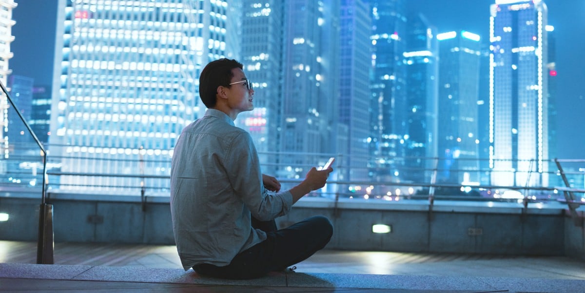 man sitting on rooftop, holding cell phone, lit up skyscrapers in front of him