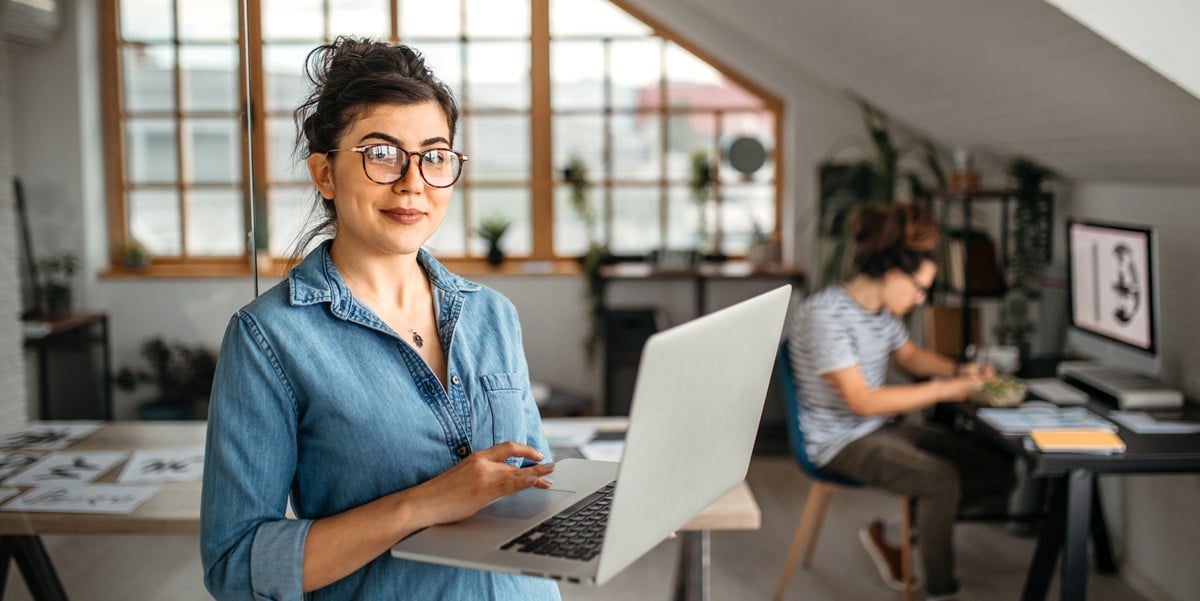 professional woman smiling, in the office, holding a laptop, colleague in the background at a desk